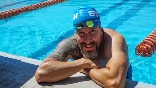 A smiling man in a blue swim cap and goggles rests on the pool edge, ready to teach how to swim laps for beginners.
