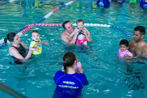 Parents and babies enjoy a swim tots class in a bright blue pool. An instructor in a blue shirt with "Aqua-Tots" visible helps a baby.