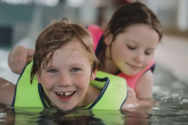 A happy boy with a missing tooth grins during 3 year old swim lessons, with a girl in a pink life vest behind him.