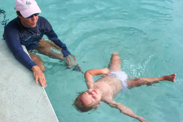 A child floats on their back in a pool, learning to swim. An instructor in a blue rash guard and pink hat watches.