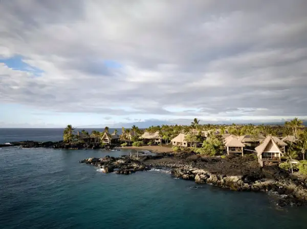 Tropical bungalows on a rocky coast in Kona, Hawaii. The calm ocean reflects the cloudy sky, a stark contrast to the power of a tsunami.