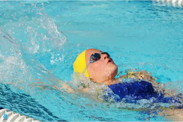 A young swimmer with a yellow cap and goggles performs the back stroke, creating a splash as they move through the blue water.