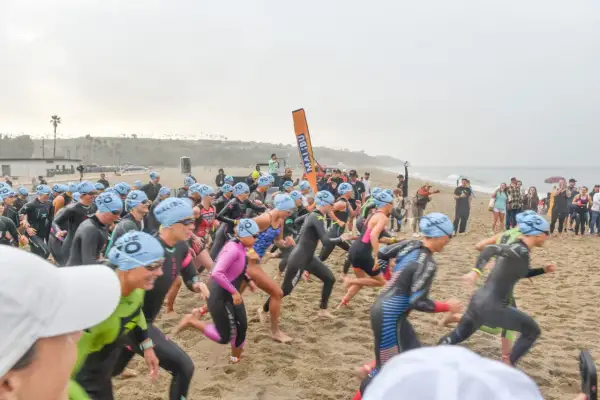 Athletes in wetsuits and blue caps sprint on the sand at the Zuma Beach Triathlon, starting their race with determination.