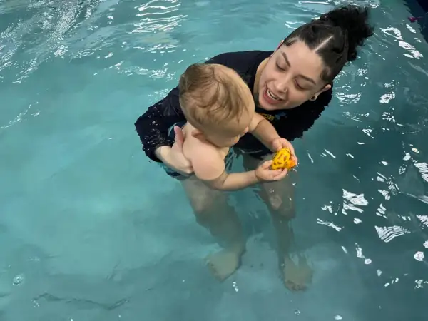A woman holds a baby in a pool, both smiling. The baby plays with a yellow toy. This scene shows that you can swim with tubes in your ears.