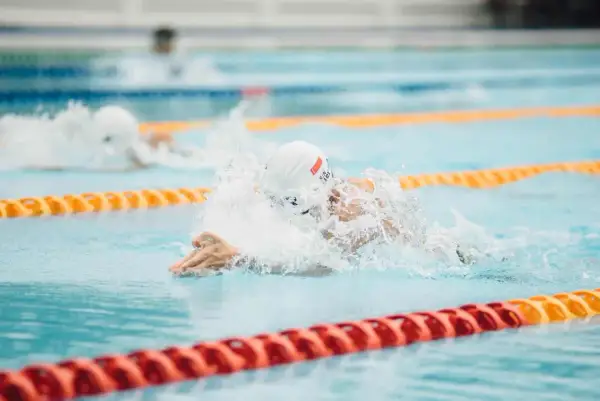 A swimmer in a white cap and goggles powers through the water, creating a splash. Their focused swimmer posture is evident as they propel forward in the lane.