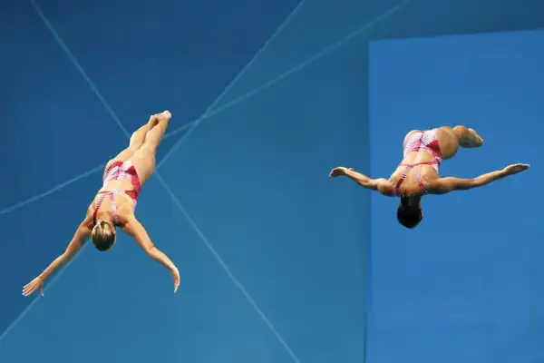Two divers perform synchronized dives, showcasing their mastery of olympic diving terms as they execute a complex maneuver against a vibrant blue background.