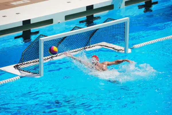 A water polo player in a red cap lunges for the ball near the net, splashing water. Intense underwater games are a thrilling spectacle.