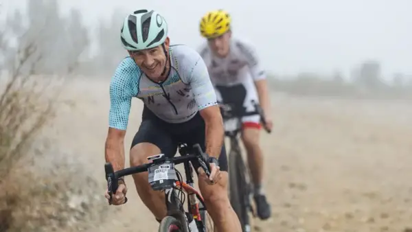 Smiles and determination on the faces of cyclists during a gravel triathlon. The lead rider, wearing a light blue and white jersey, grins as he navigates the dusty course.