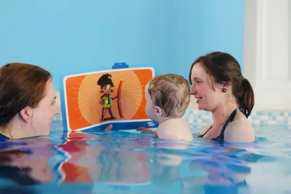 A toddler enjoys a fun toddler swim school lesson, looking at a colorful book with two smiling instructors in the pool.