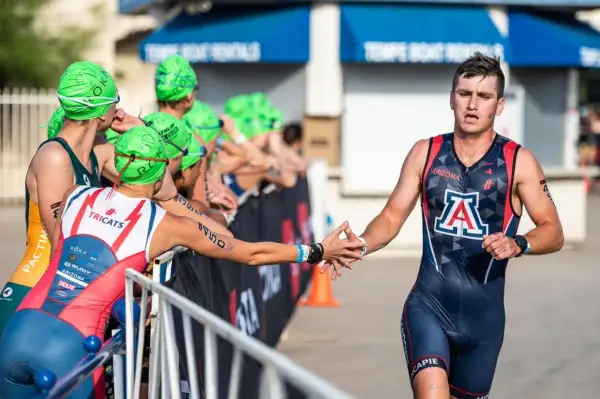 A male NCAA triathlon athlete in a dark blue and red suit with an "A" logo high-fives a competitor.