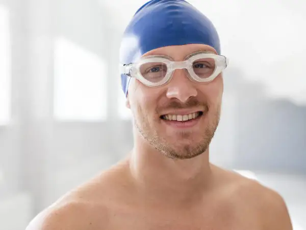 A smiling man in a blue swim cap and goggles, ready to swim. He's wondering, "When can I swim after wisdom teeth removal?