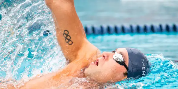 Swimmer in backstroke position, with Olympic rings tattoo on arm, mid-stroke, water splashing.
