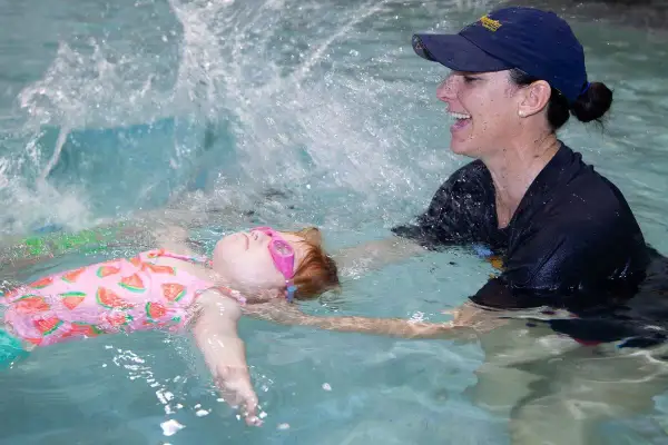 A child in a watermelon swimsuit and pink goggles floats on their back while a smiling instructor helps them practice treading water.