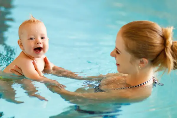 A happy baby laughs during baby and me swim classes, held by their mother in a bright blue pool.