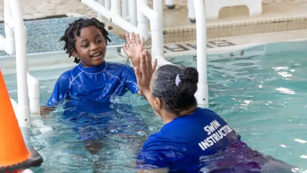 A young boy with dreadlocks in a blue rash guard gives a high-five to a swim instructor. This is a moment of connection during an autism swim lesson.