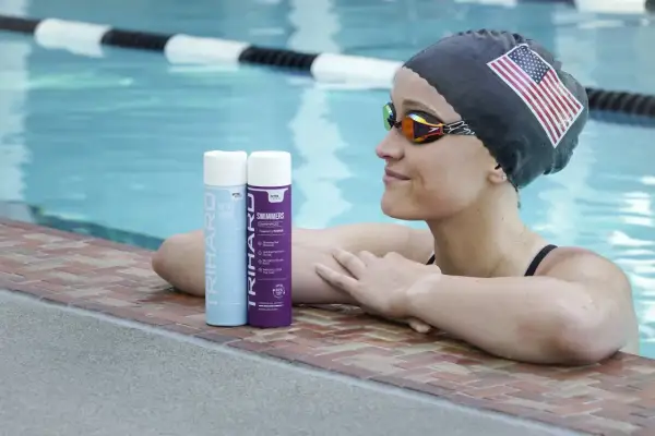 Swimmer with an American flag cap and goggles rests by the pool, with TRIHARD swimmer's shampoo bottles nearby, offering a solution on how to protect skin from chlorine.