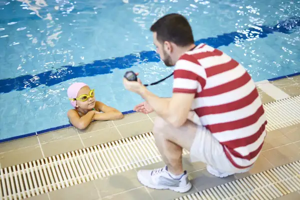 A mobile swim instructor crouches by the pool, holding a stopwatch, as a young girl in a pink cap and goggles rests her arms on the edge.