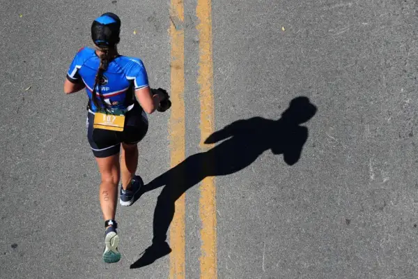 A runner in blue triathlon apparel, bib number 107, runs on a road, casting a long shadow.