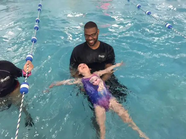 A smiling instructor helps a young girl in a Frozen swimsuit learn how to float on her back in a swimming pool.