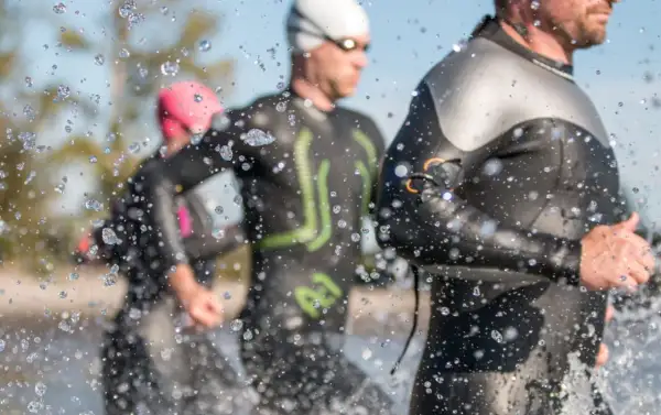 Triathletes in Ironman wetsuits run through water, splashes obscuring the view. Following Ironman wetsuit rules, they prepare for the race.