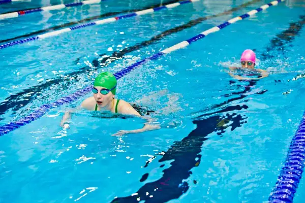 Two swimmers in a pool, one in a green cap doing breaststroke, the other in a pink cap swimming freestyle.