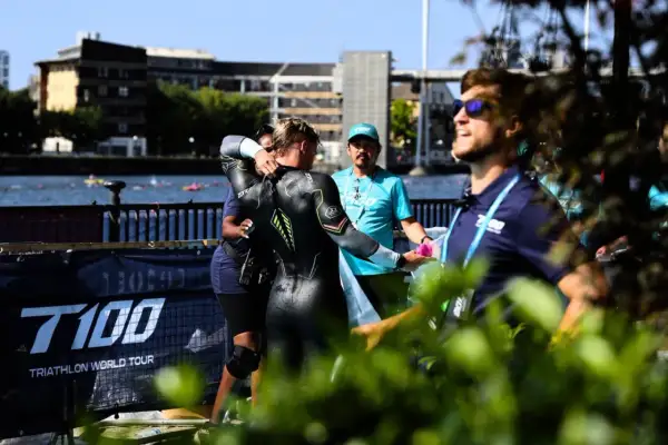 A triathlete in a wetsuit is helped by staff during a triathlong transition. The T100 Triathlon World Tour banner is visible.